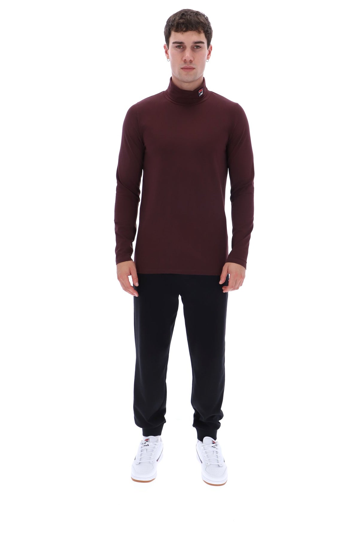 A man modeling a long-sleeve mock neck turtleneck sweater in burgundy, paired with black pants and white sneakers, against a solid white background.