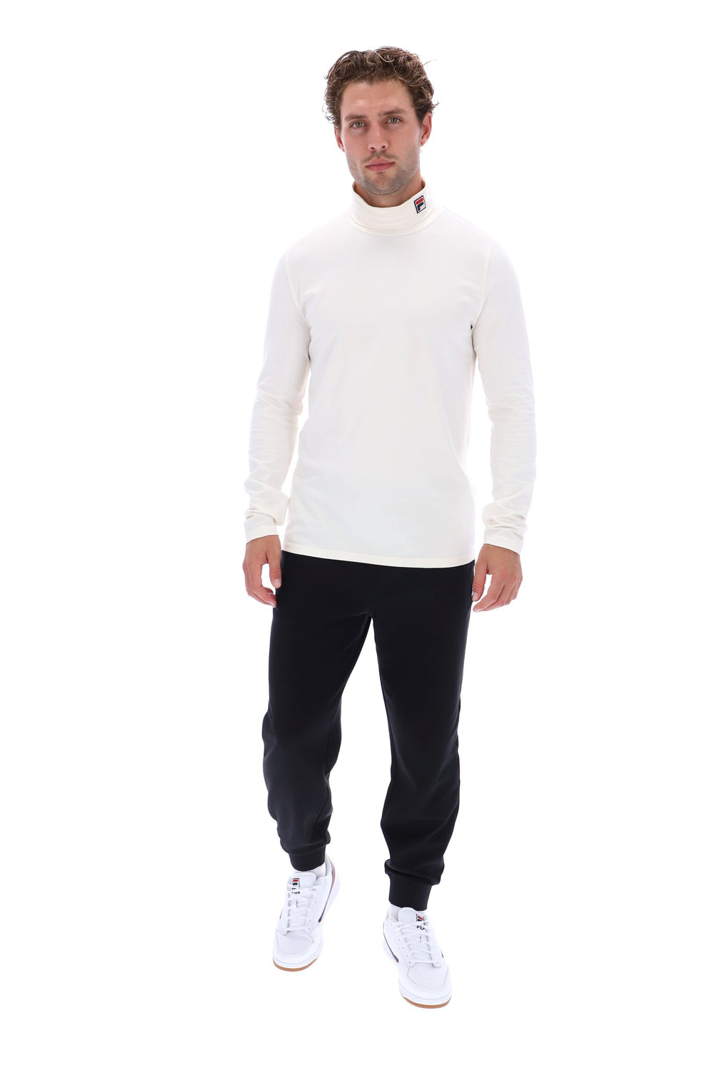 A man modeling a white long-sleeve mock neck sweater, black jogger pants, and white sneakers against a solid white background.