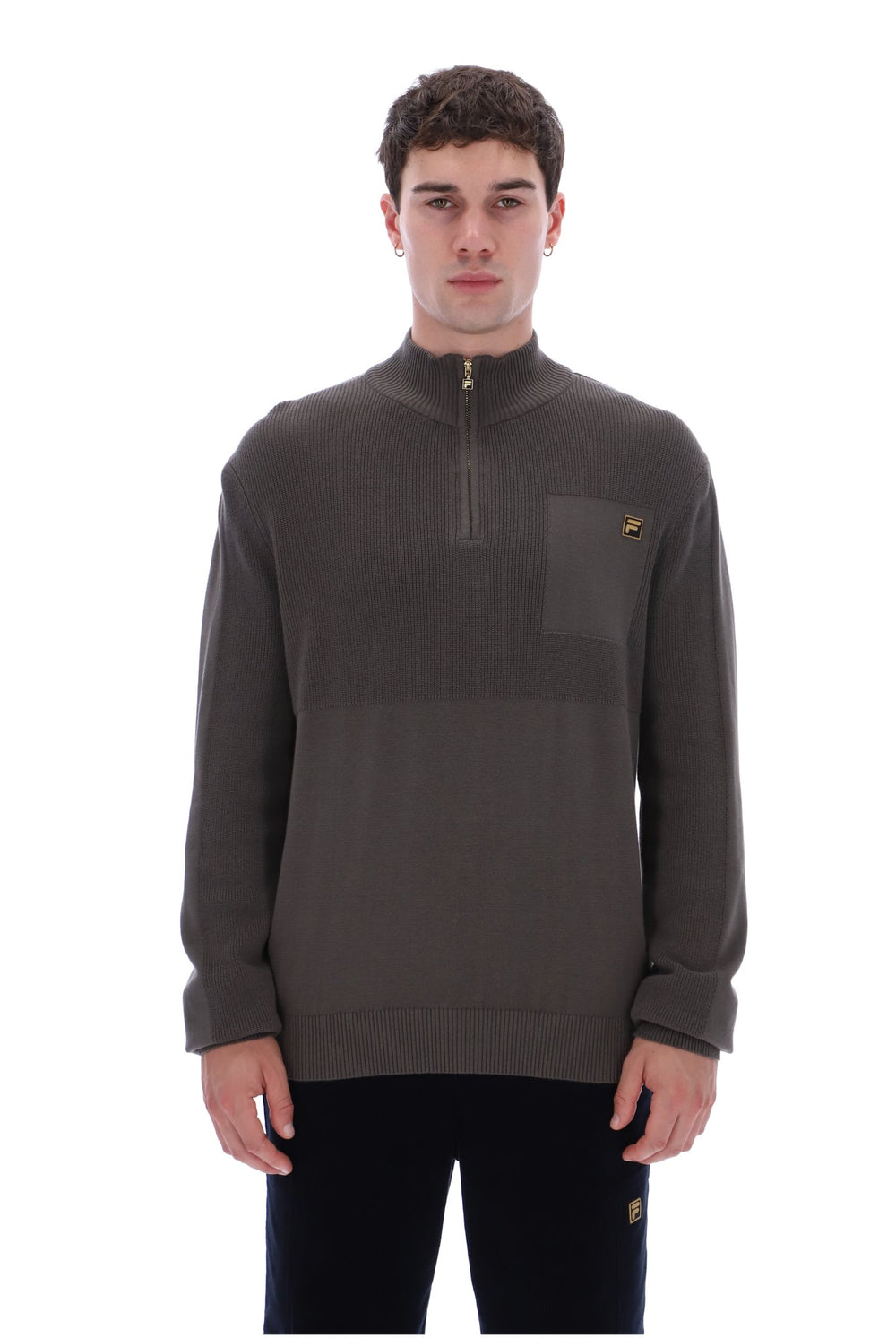 A man wearing a dark brown quarter-zip pullover sweater with a chest pocket and a small gold embroidered logo, paired with black pants, against a plain white background.