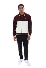 A man modeling a color-block zip-up jacket with dark maroon sleeves and front, and an off-white chest panel, paired with black pants and white sneakers, on a solid white background.