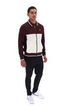 A man modeling a color-blocked zip-up track jacket in maroon and cream, paired with black track pants and white sneakers, against a plain white background.