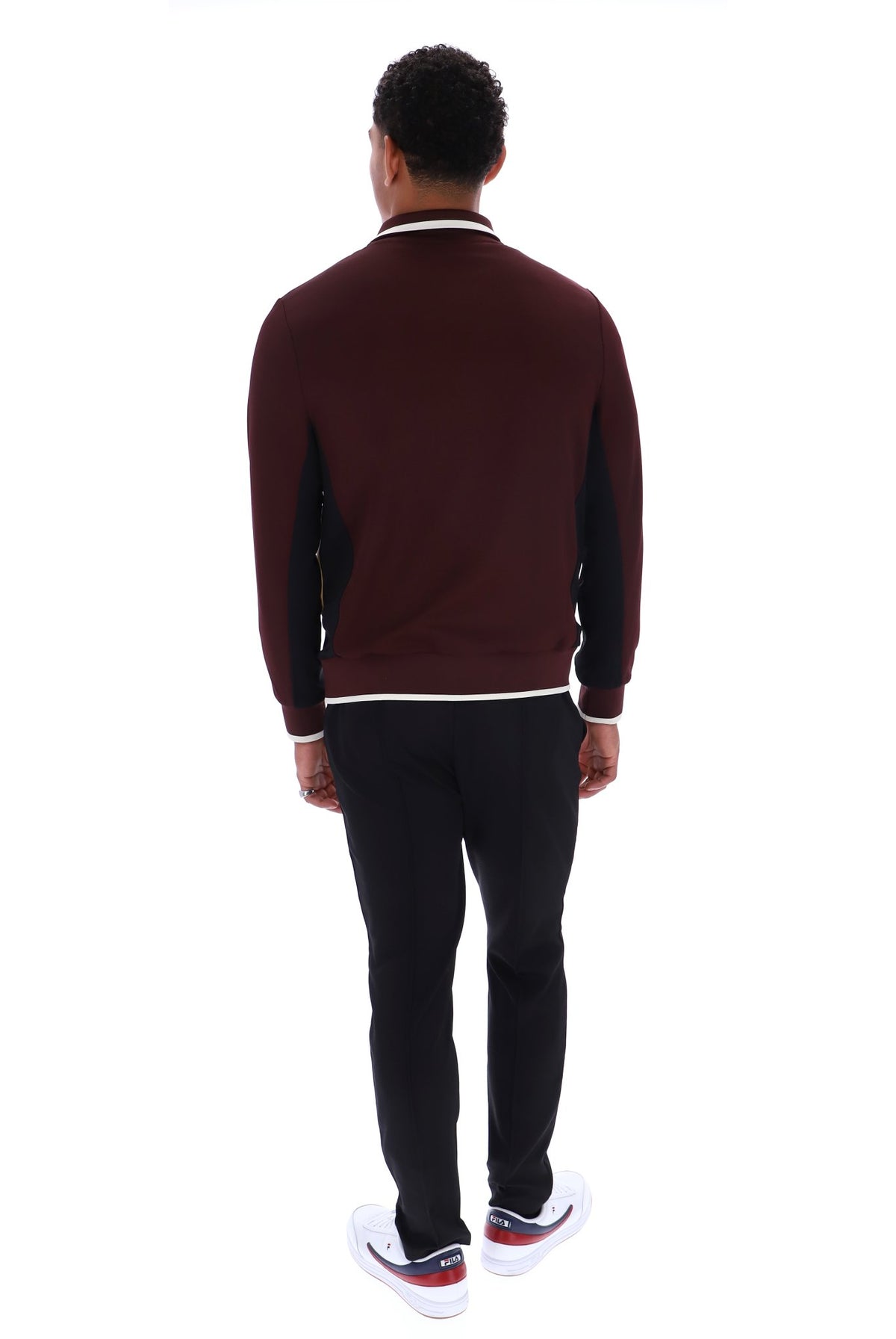 A man viewed from behind wearing a burgundy and black long-sleeve knit sweater with white trim, black pants, and white sneakers, shot against a plain white background.