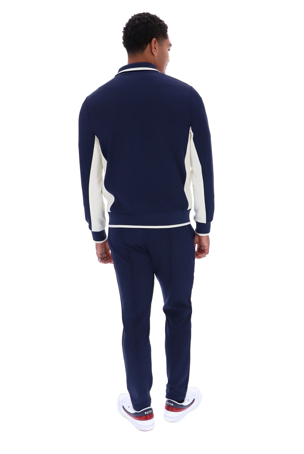 A man viewed from the back wearing a navy blue knit tracksuit with white contrast panels and white sneakers, set against a plain white background.