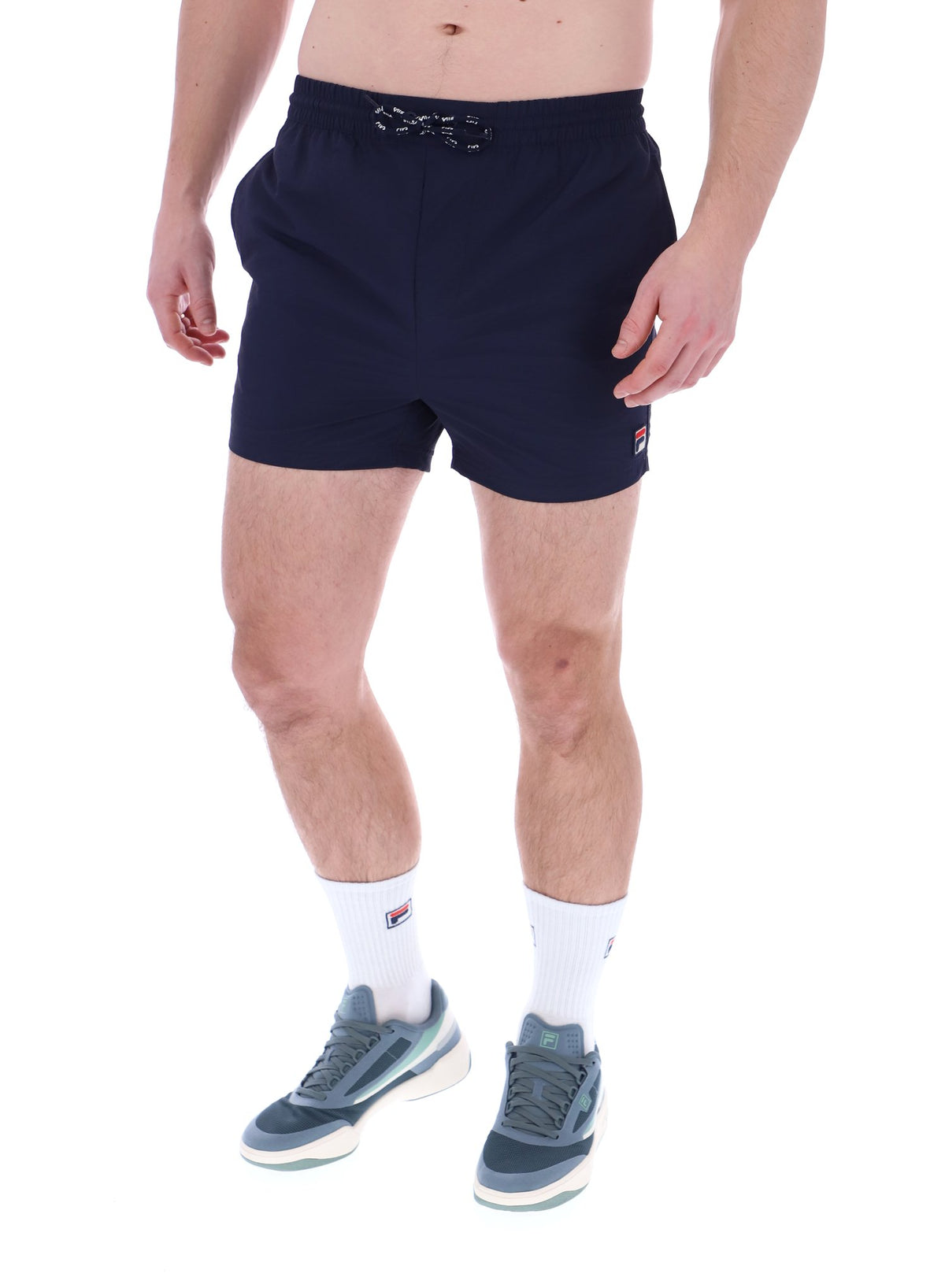 A person wearing black Artoni Swim Shorts with a drawstring waistband and the FILA logo on the leg, paired with white socks and grey sneakers.