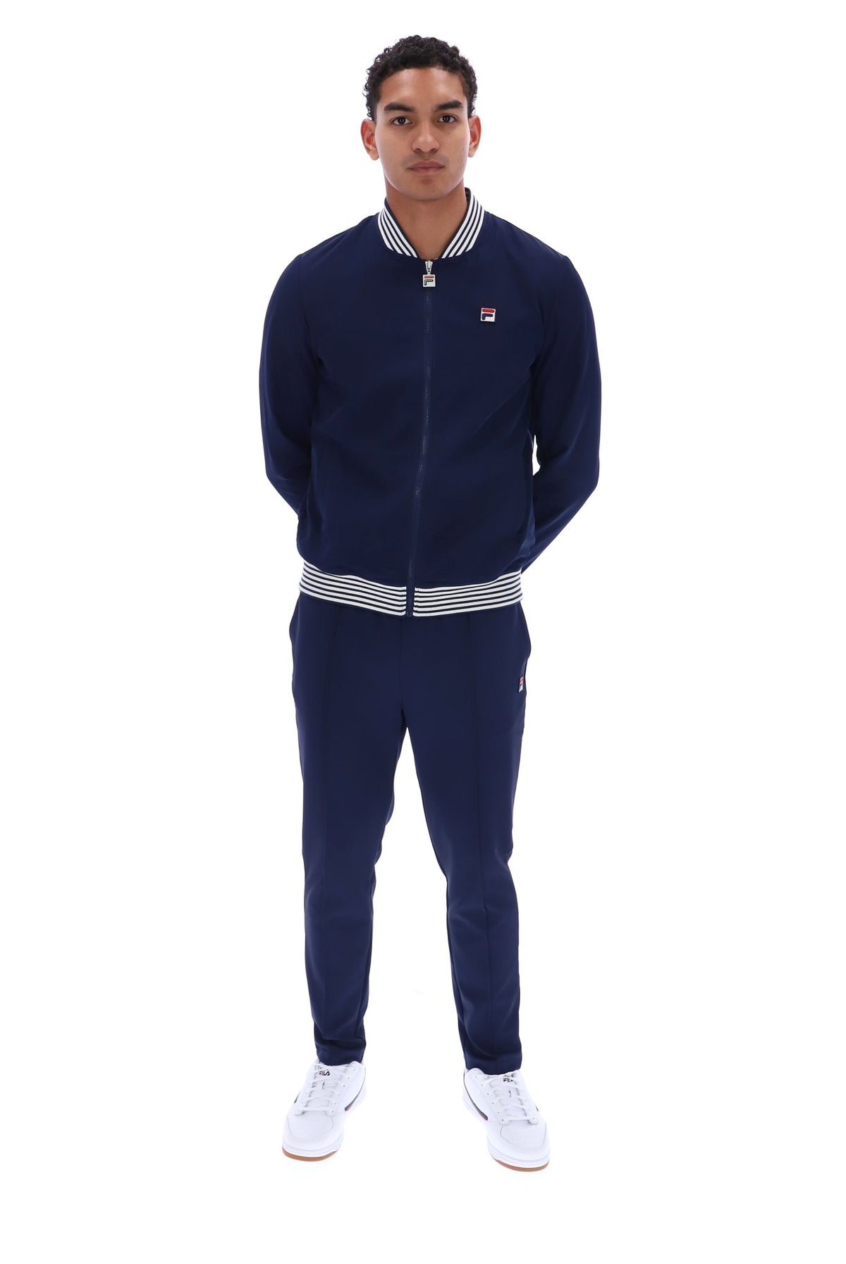 A man modeling a navy blue Fila tracksuit, featuring a zip-up jacket with white and navy striped ribbed collar, cuffs, and hem, paired with matching navy sweatpants and white sneakers, shot against a solid white background.