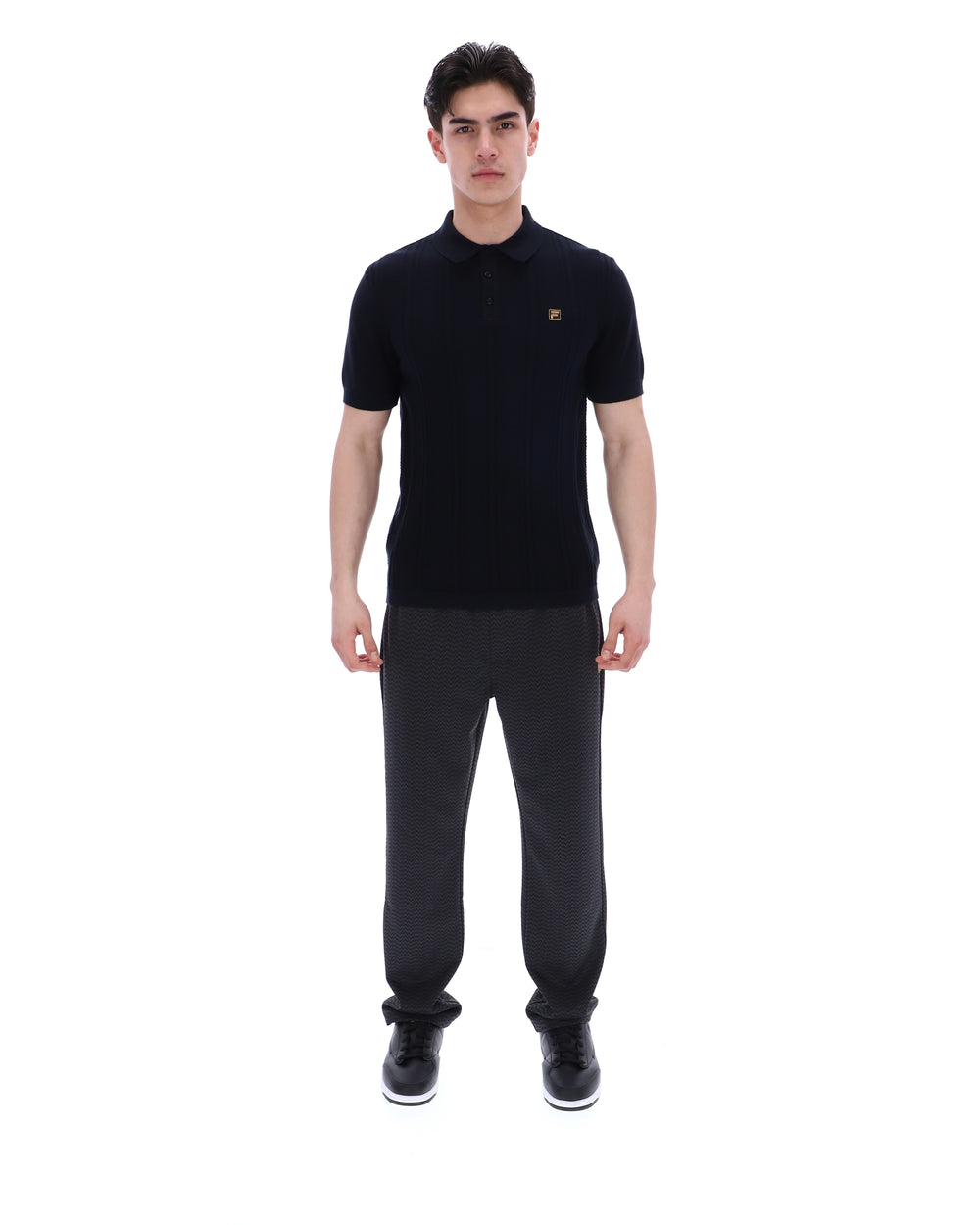Man wearing a black polo shirt with a small FILA logo on the chest, paired with black FILA joggers, against a white background