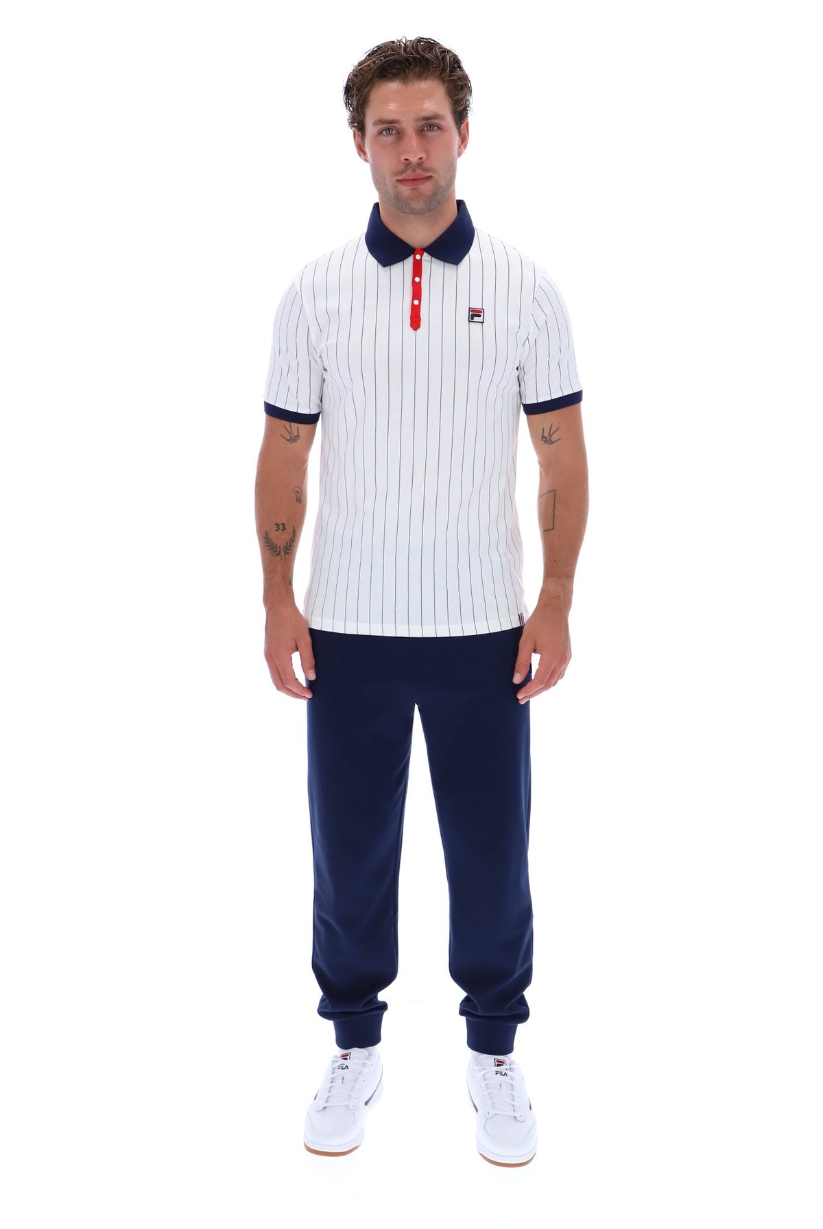 A man modeling a white short-sleeve polo shirt with navy blue pinstripes, a navy collar, and a red button placket, paired with navy pants and white sneakers, against a plain white background.