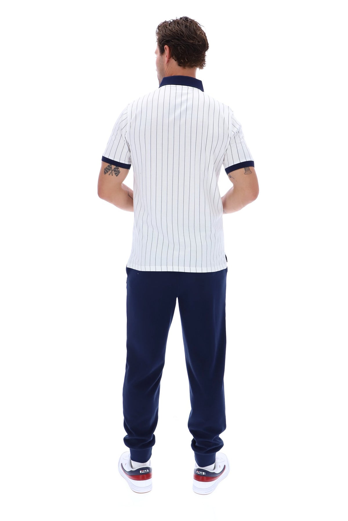 A man viewed from behind wearing a white short-sleeve polo shirt with thin navy vertical pinstripes and navy trim on the collar and sleeve cuffs, paired with navy jogger pants and white sneakers.