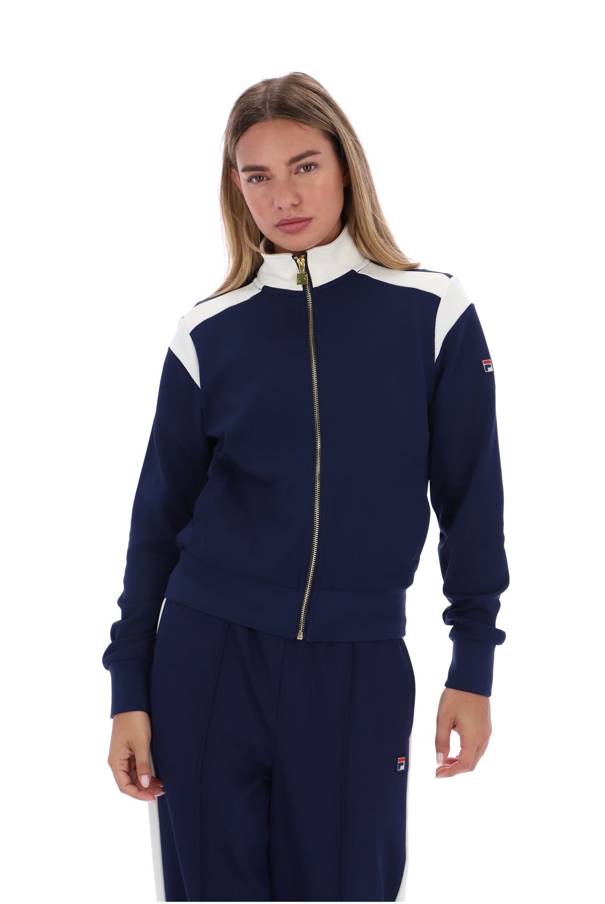 A woman modeling a navy blue and white track jacket with a gold zipper, paired with matching navy pants, against a plain white background.