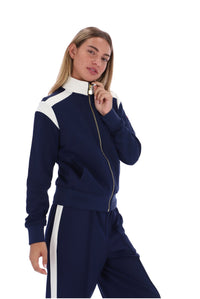 A woman modeling a navy blue and white athletic tracksuit with a gold zipper, featuring a zip-up jacket and matching pants with white side stripes, set against a plain white background.