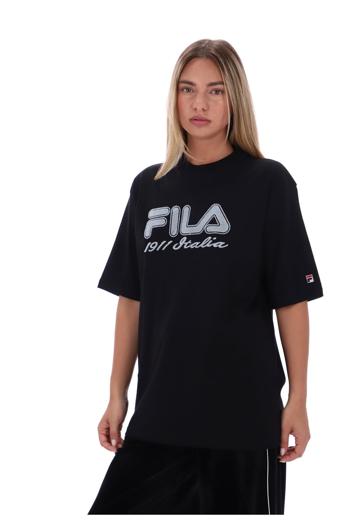 A woman modeling an oversized black FILA t-shirt with a large white and gray logo on the chest and a small FILA logo on the sleeve, set against a plain white background.