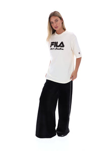 A woman modeling a white FILA All Asta graphic t-shirt with black lettering, paired with black wide-leg pants, against a solid white background.