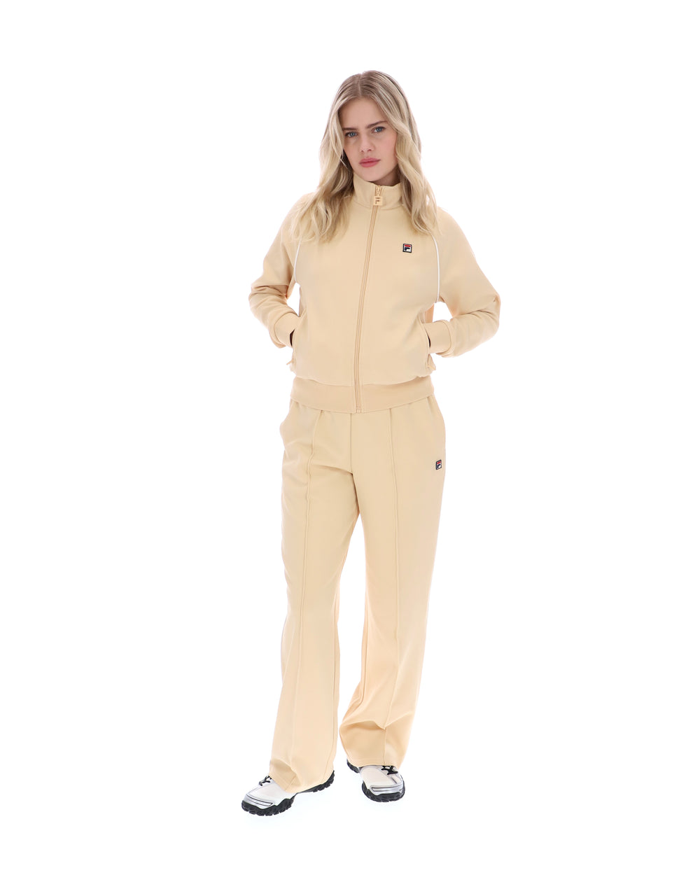 Woman wearing a beige track jacket with a FILA logo on the chest, paired with matching bottoms, against a white background