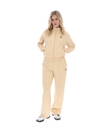 Woman wearing a beige track jacket with a FILA logo on the chest, paired with matching bottoms, against a white background