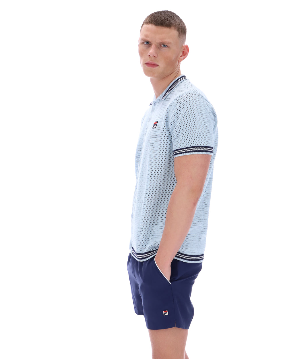 Man wearing a light blue patterned polo shirt with a FILA log on the chest and navy shorts, against a white background.