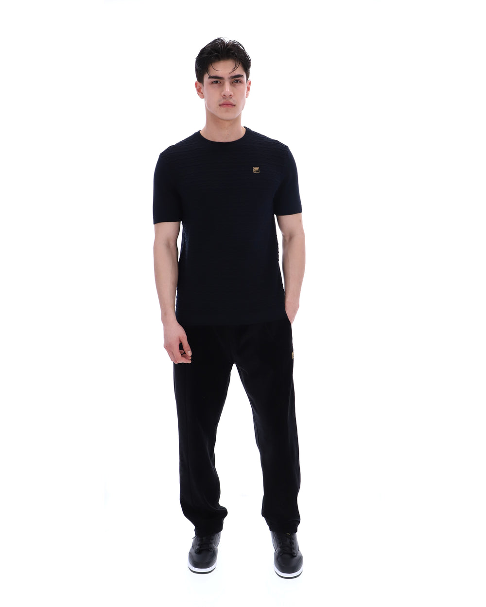 Man wearing a black t-shirt with a small FILA Gold logo on the chest with matching bottoms on a white background