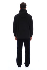 A man viewed from the back wearing a black hooded sweatshirt and black pants against a plain white studio background.