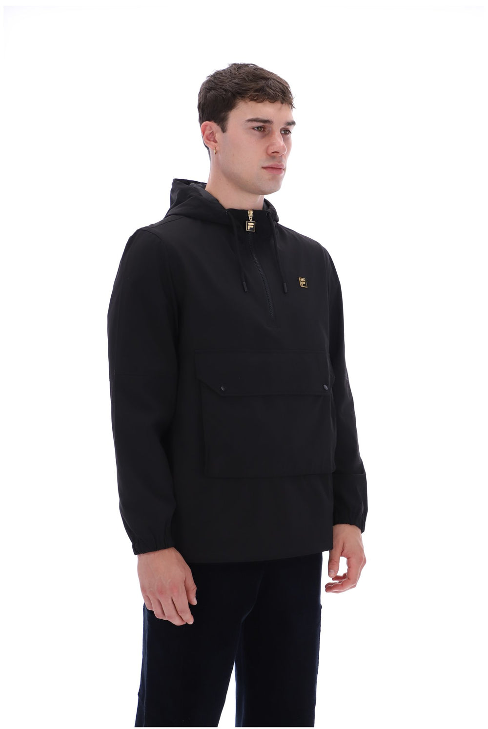 A man wearing a black hooded pullover jacket with a small gold embroidered logo on the left chest, paired with black pants, photographed against a solid white background.