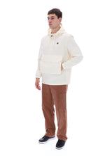 A man modeling a cream-colored hooded pullover jacket with a front flap pocket, paired with brown pants and black sneakers, against a solid white background.