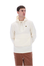 A man wearing a cream-colored pullover hoodie with a kangaroo pocket and brown pants, standing against a solid white background.