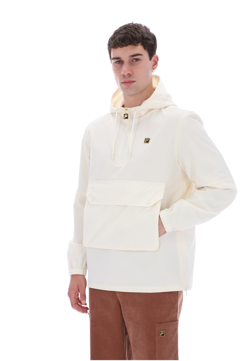 A man modeling a cream-colored hooded windbreaker jacket with a large front flap pocket, paired with brown pants, against a solid white background.