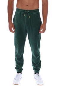 A man wearing dark green velvet jogger sweatpants with a drawstring waist and white sneakers, shown from the chest down against a plain white background.