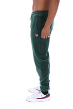 A side profile view of a person wearing dark green Fila jogger sweatpants and white sneakers with red and navy accents, set against a plain white background.