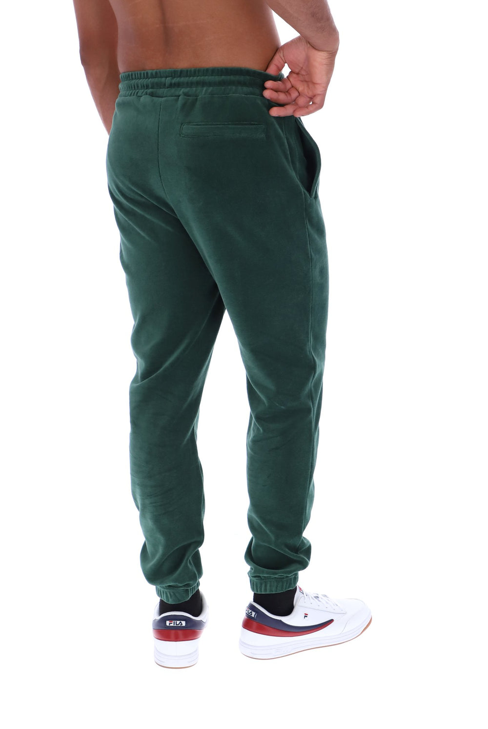 A man wearing dark green corduroy jogger pants, shown from the back. He is wearing white sneakers with red and navy accents and black socks, against a plain white background.