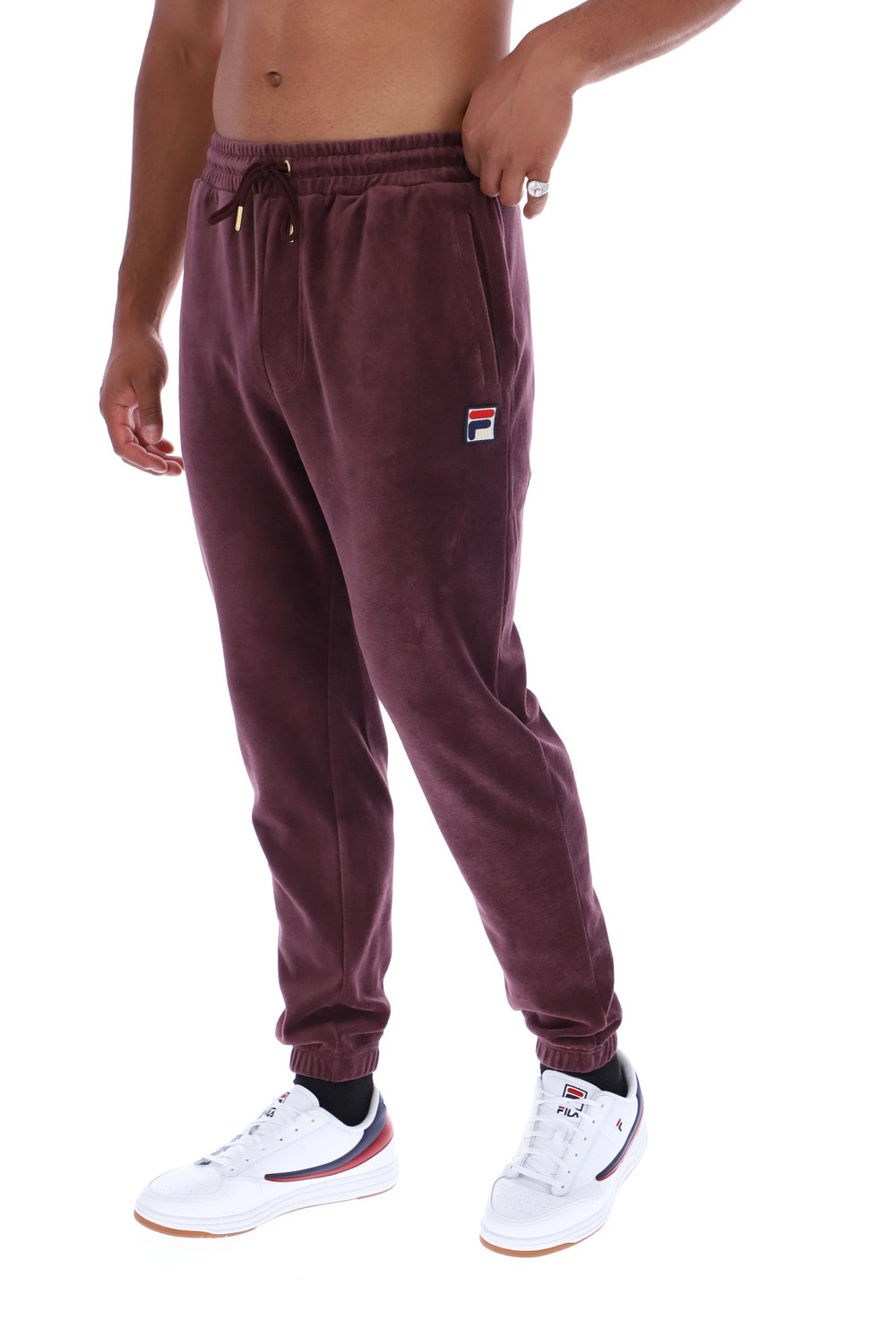 A person wearing burgundy fleece jogger sweatpants with a small Fila logo patch on the left thigh, paired with white sneakers, shown against a plain white background.