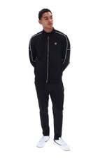 A man modeling a black FILA tracksuit with white piping details and a gold zipper, paired with white sneakers, on a solid white background.