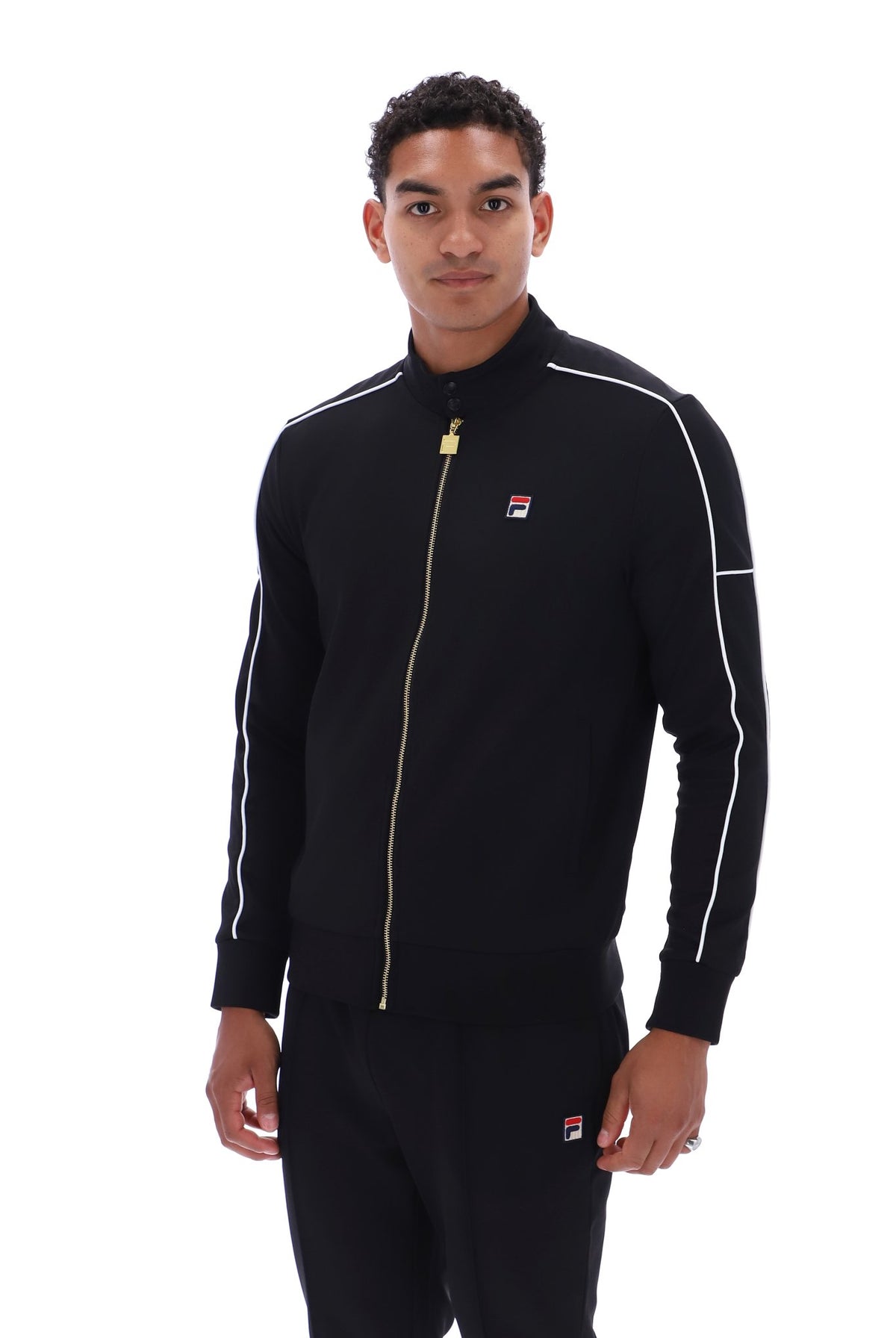 A man wearing a black Fila track jacket with white piping and a gold zipper, paired with matching black track pants, against a plain white background.