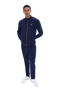 A man modeling a navy blue tracksuit with light blue piping details, featuring a zip-up jacket and matching pants, paired with white sneakers, against a solid white background.