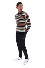 A man wearing a long-sleeve crewneck sweater with horizontal stripes in black, beige, and white, paired with black sweatpants and white sneakers, standing against a plain white background.