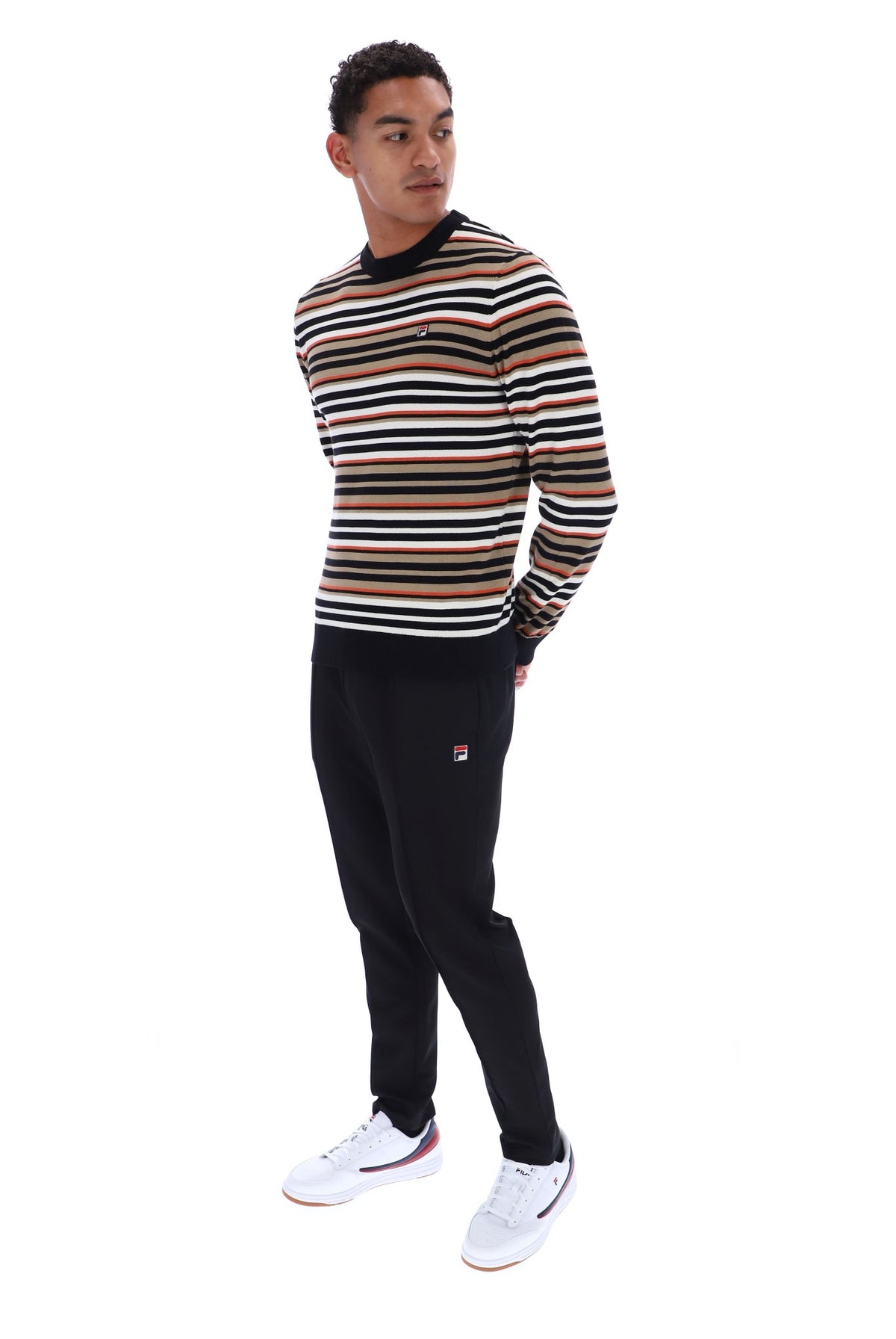 A man wearing a long-sleeve crewneck sweater with horizontal stripes in black, beige, and white, paired with black sweatpants and white sneakers, standing against a plain white background.