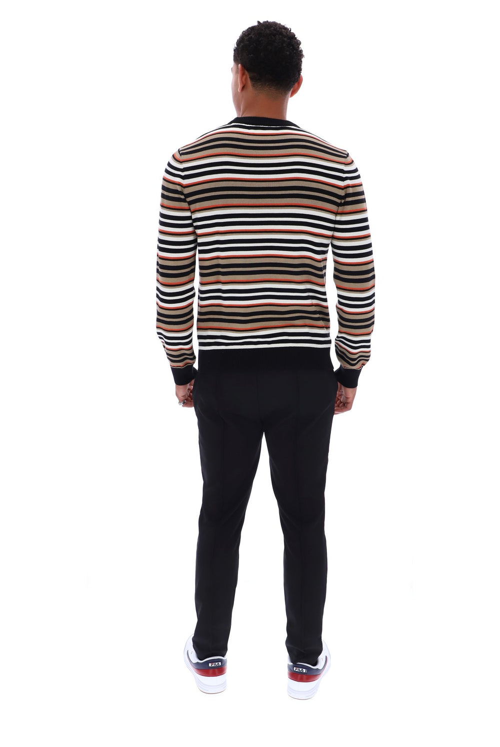 A man viewed from behind wearing a long-sleeve crewneck sweater with horizontal stripes in black, brown, beige, and white, paired with black pants and white sneakers.