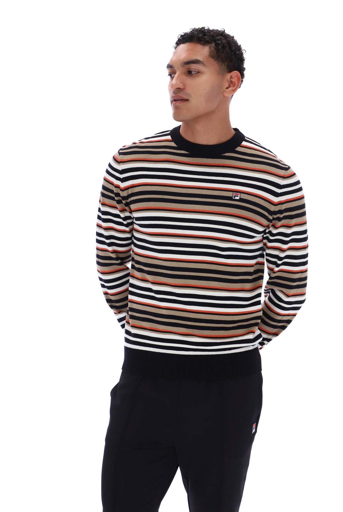 A man wearing a long-sleeve crewneck sweater with horizontal stripes in black, white, and tan, paired with black sweatpants, photographed against a solid white background.