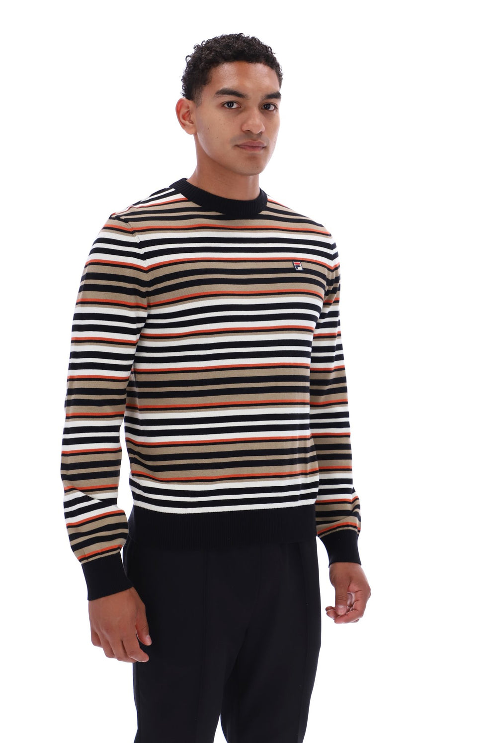 A man wearing a long-sleeve crewneck sweater with horizontal stripes in black, white, tan, and brown, paired with black pants, photographed against a plain white background.