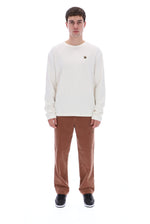 A man modeling a cream long-sleeve crewneck sweatshirt with a small brown logo on the left chest, paired with brown corduroy pants and black sneakers, against a solid white background.