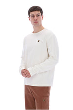 A man wearing a cream-colored long-sleeve crewneck sweatshirt with a small brown logo on the left chest, paired with brown pants, against a solid white background.