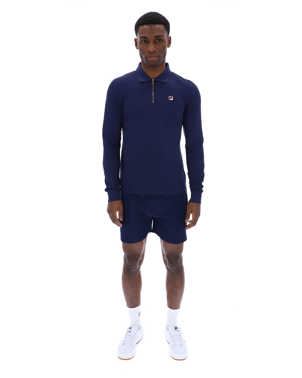 Man wearing a navy blue zip-up collared long-sleeve with a FILA logo on the chest, paired with matching shorts, on a white background