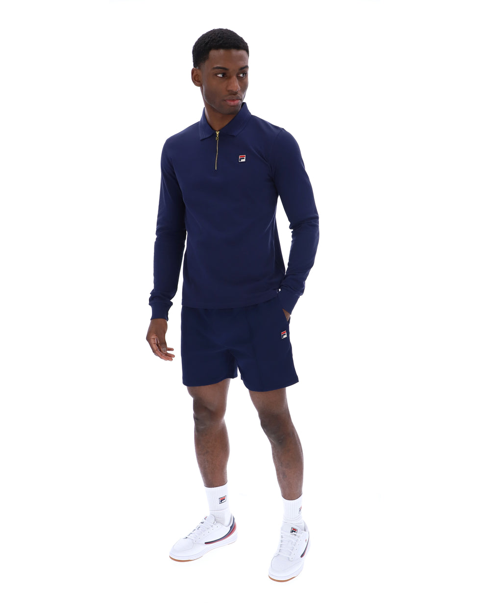 Man wearing a navy blue zip-up collared long-sleeve with a FILA logo on the chest, paired with matching shorts, on a white background