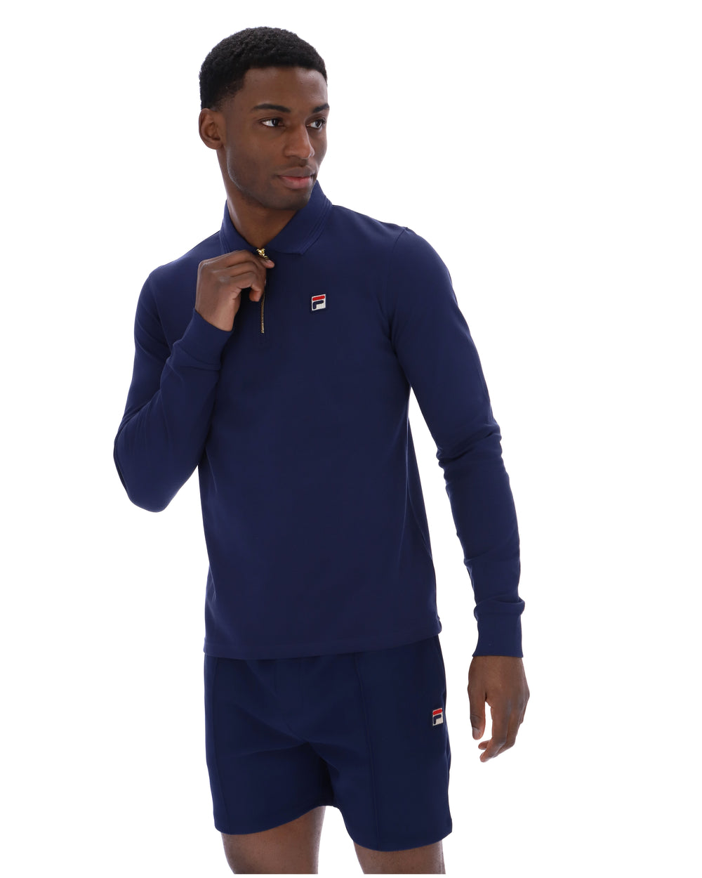 Man wearing a navy blue zip-up collared long-sleeve with a FILA logo on the chest, paired with matching shorts, on a white background