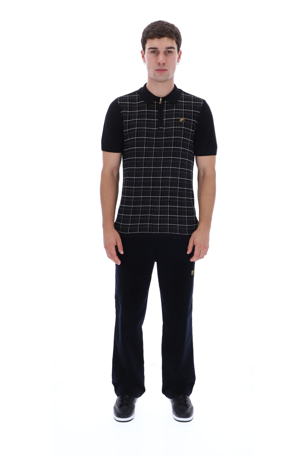 A man modeling a short-sleeve polo shirt with a black and white windowpane plaid pattern on the front and solid black sleeves, paired with black trousers and black sneakers, against a plain white background.