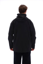 A person viewed from the back wearing a black hooded jacket and dark pants against a plain white studio background.