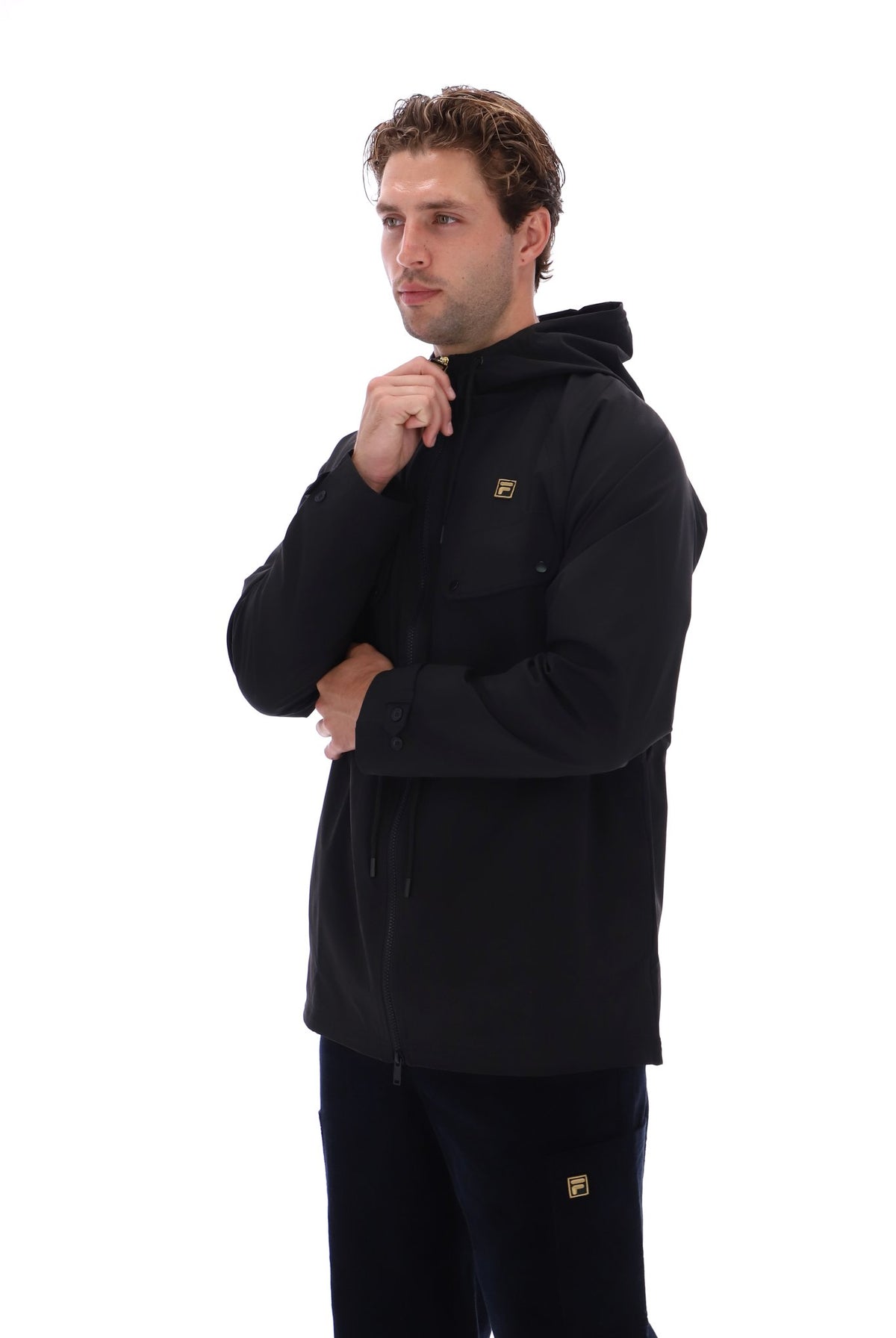 A man wearing a black hooded jacket with a small gold logo on the chest, paired with black pants, photographed against a solid white background.