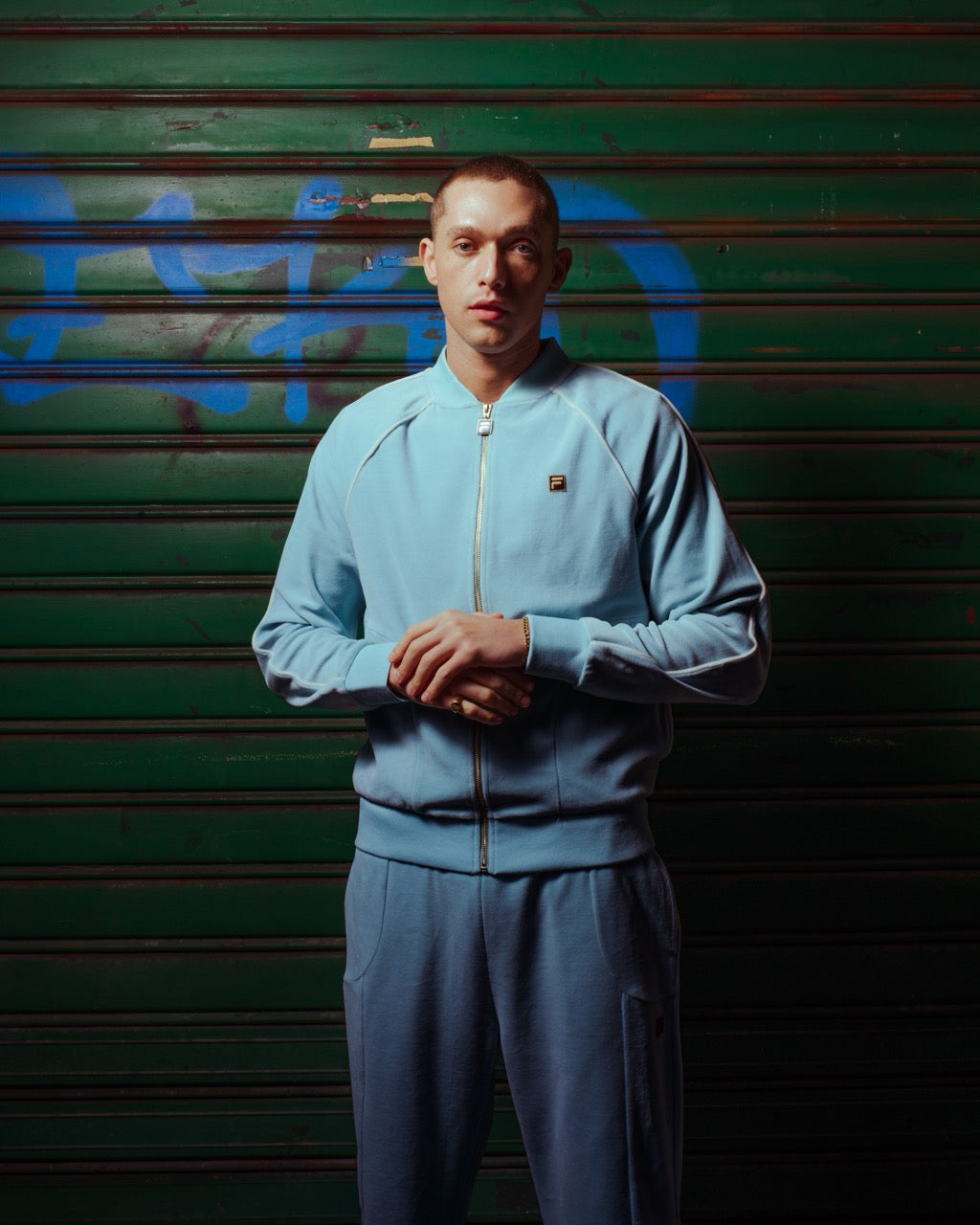 Person wearing a light blue tracksuit standing in front of a green shutter with graffiti.