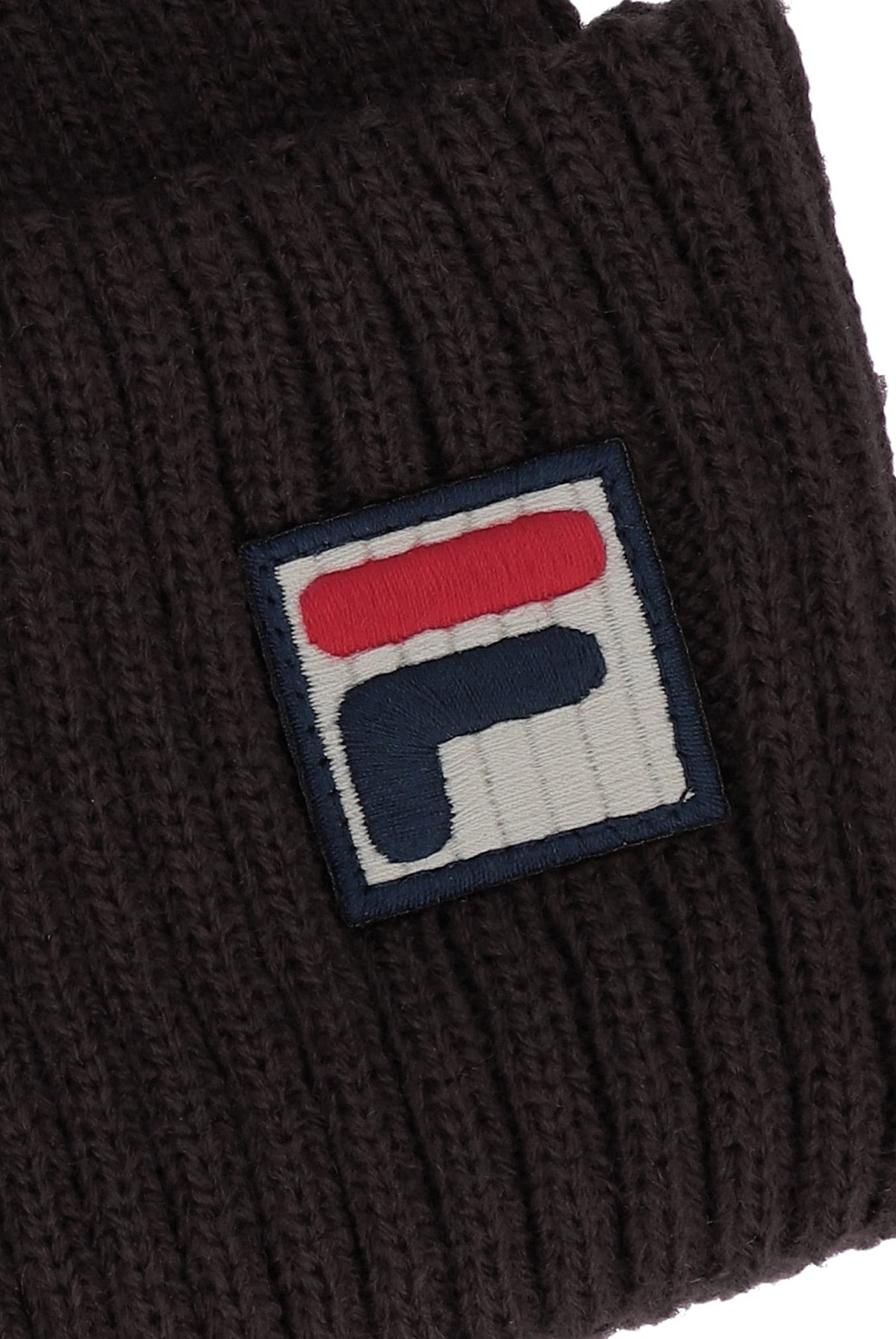 Close-up of a dark brown ribbed knit beanie featuring a square embroidered patch with the red, white, and navy blue FILA logo.