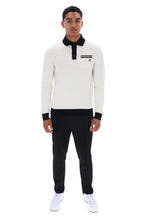 A man modeling a long-sleeve knit polo shirt in off-white with black collar, cuffs, and placket, paired with black trousers and white sneakers, shot against a plain white background.