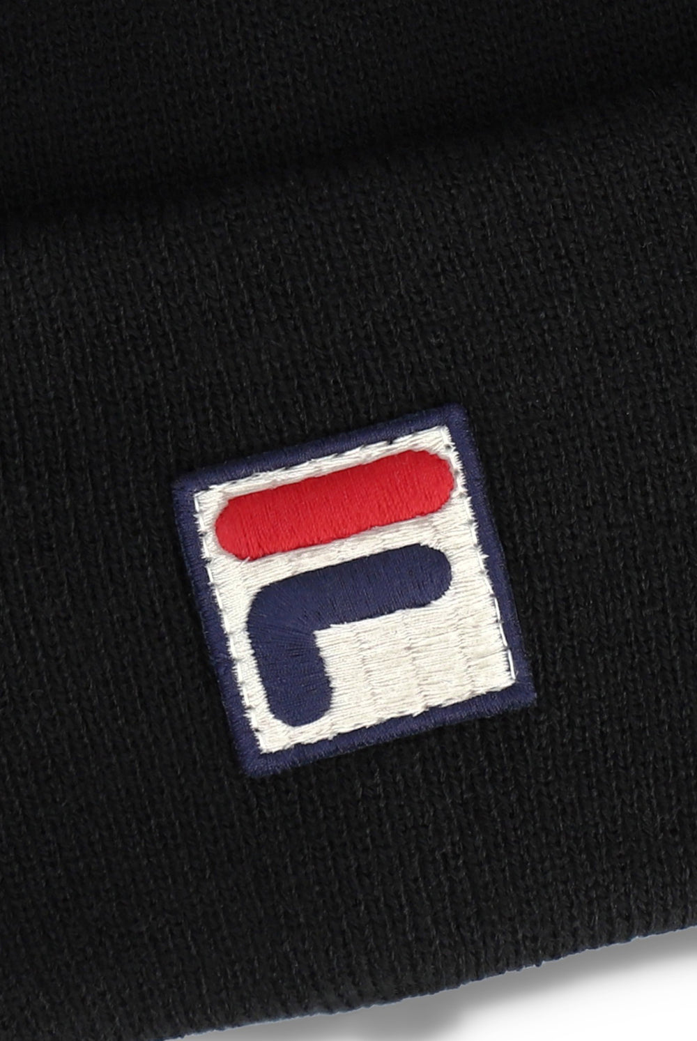 Close-up of a black knit beanie featuring an embroidered square patch with the Fila logo in red, white, and navy blue.