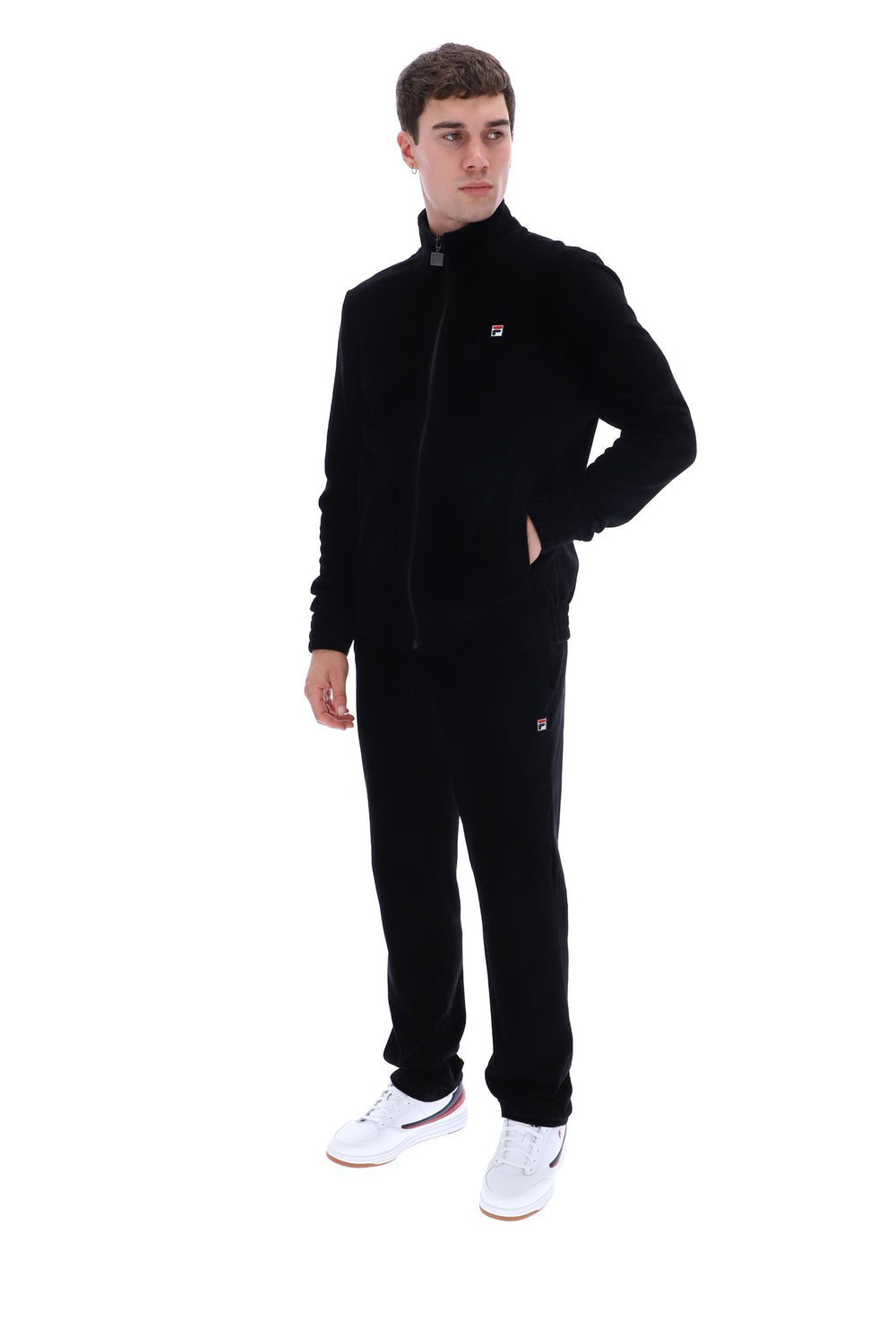 A young man modeling a black fleece tracksuit with a zip-up jacket and matching sweatpants, featuring a small red and white logo on the chest and thigh, paired with white sneakers, against a plain white background.
